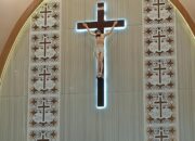 Tampak sebuah altar dengan salib besar dan patung Yesus Kristus yang disinari cahaya biru lembut. Di sekelilingnya terdapat panel dekoratif bermotif salib dan burung merpati, melambangkan iman dan perdamaian. Foto: Istimewa.