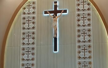 Tampak sebuah altar dengan salib besar dan patung Yesus Kristus yang disinari cahaya biru lembut. Di sekelilingnya terdapat panel dekoratif bermotif salib dan burung merpati, melambangkan iman dan perdamaian. Foto: Istimewa.