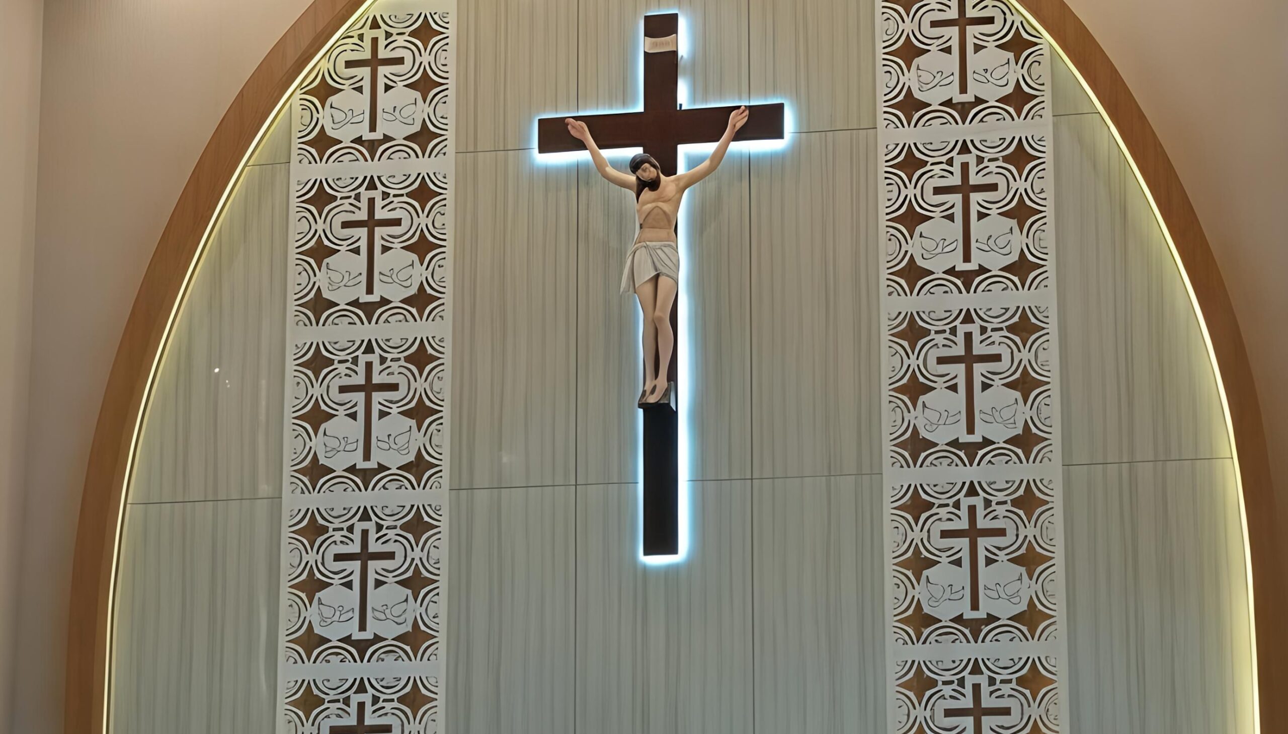 Tampak sebuah altar dengan salib besar dan patung Yesus Kristus yang disinari cahaya biru lembut. Di sekelilingnya terdapat panel dekoratif bermotif salib dan burung merpati, melambangkan iman dan perdamaian. Foto: Istimewa.